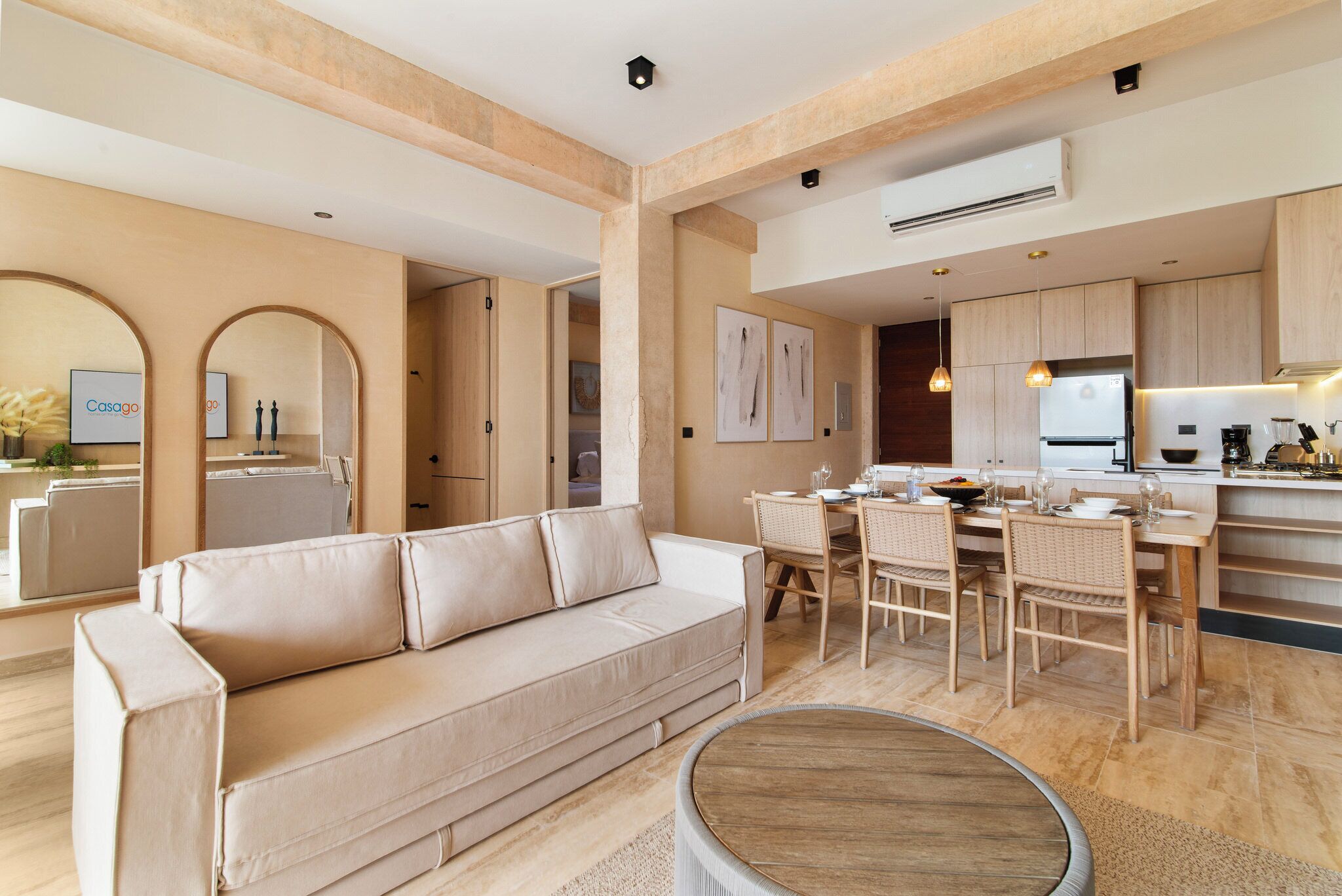 A modern living room with a beige sofa, dining area with wooden chairs, and a kitchen with light wood cabinets and a white refrigerator.