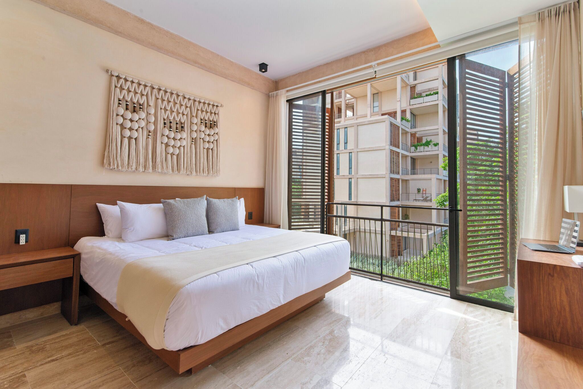 Una habitación de hotel moderna con una cama grande, cabecera de madera, un tapiz de macramé en la pared, un balcón con vistas a los balcones del edificio y un escritorio con una lámpara.