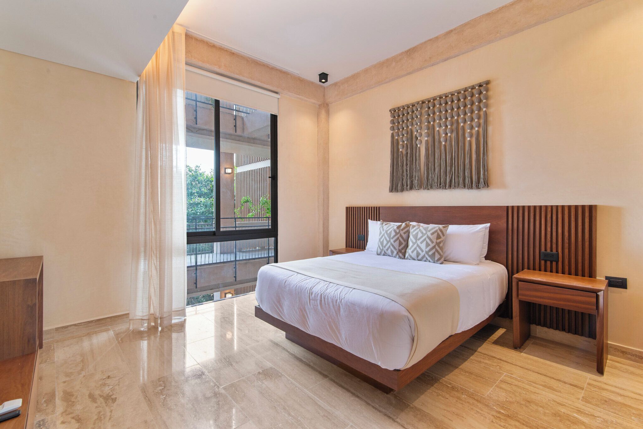 A modern hotel room with a large bed, a wooden headboard, a nightstand, and a view of the outdoors through a sliding door.