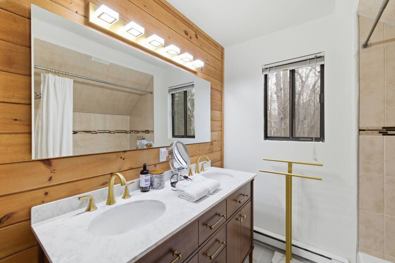A bathroom with a wooden vanity, two sinks with gold faucets, a large mirror, and a window with a view of trees.