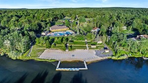 An aerial view of a lakeside property with a dock, swimming area, and residential buildings surrounded by dense forest. - Lake Monster Family Adventure! Kayak fishing Firepit Hot tub Sauna Games & More (Tobyhanna)
