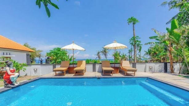 A swimming pool with lounge chairs and umbrellas, surrounded by tropical plants and a clear sky. - Kalpataru Cottage (Bali)