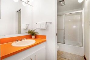 Casa de banho com lavatório branco, bancada laranja e cabine de duche em vidro.