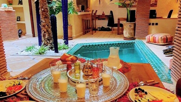 A dining area with a pool, featuring a table set with plates, glasses, and a pitcher, surrounded by potted plants and a decorative ceiling.