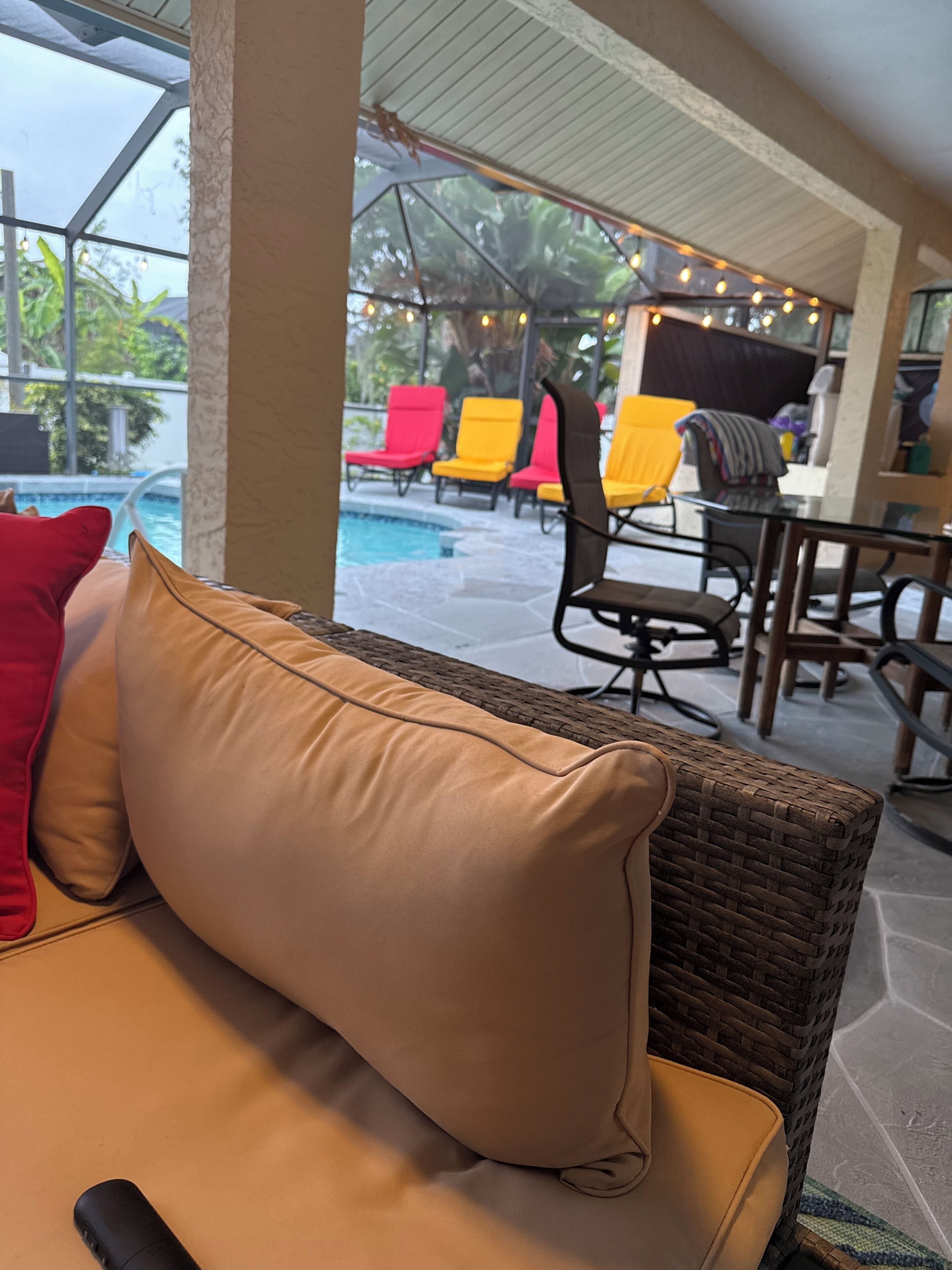 A wicker sofa with a beige cushion and a red pillow, a glass table, and a pool area with colorful lounge chairs.