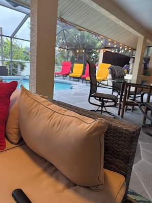A wicker sofa with a beige cushion and a red pillow, a glass table, and a pool area with colorful lounge chairs.