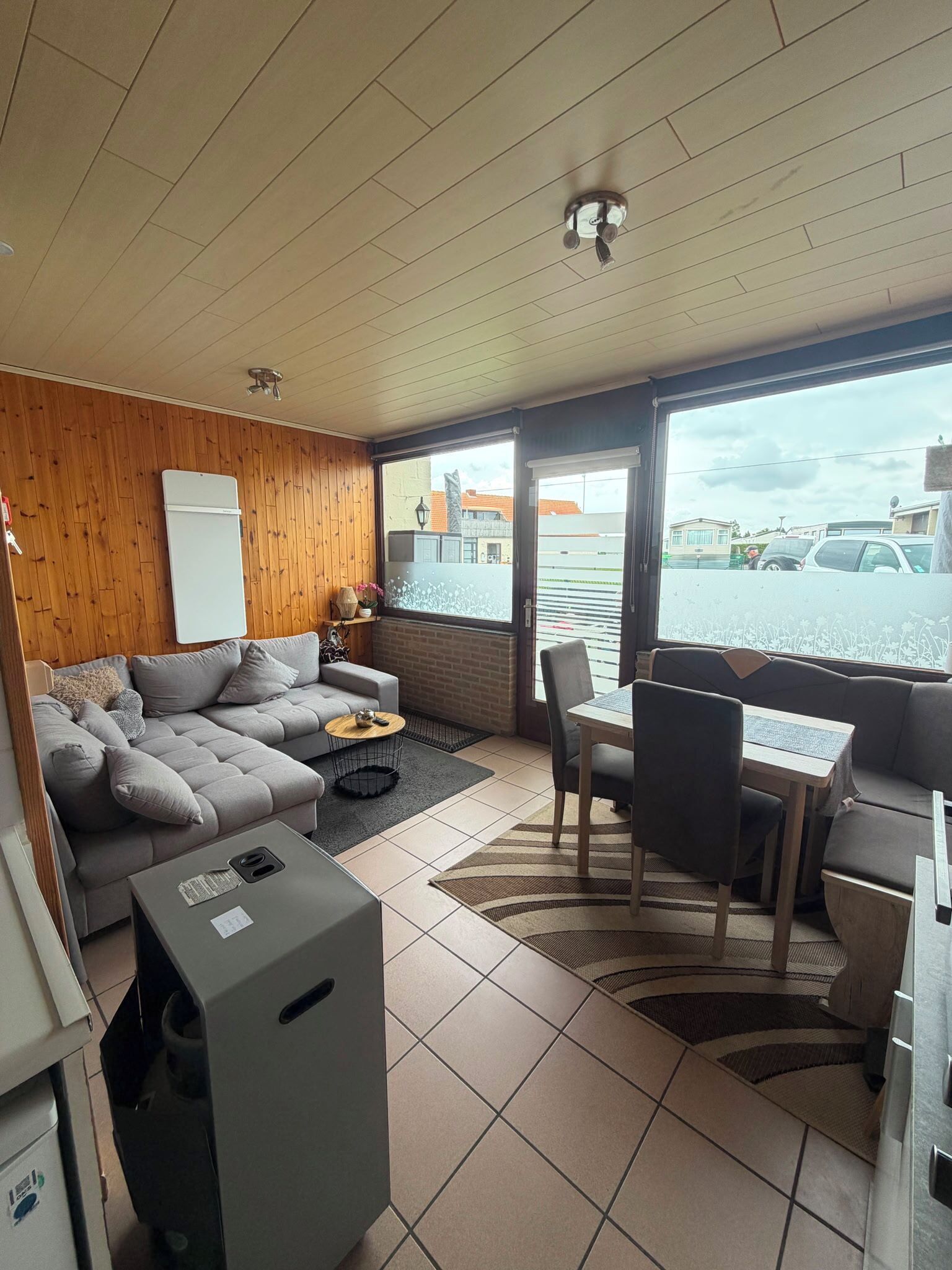 A room with a sofa, a dining table with chairs, a small kitchen area, and a view of the outside.