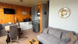 A modern living room with a grey sofa, a wooden dining table, and a kitchen area with a TV and refrigerator.