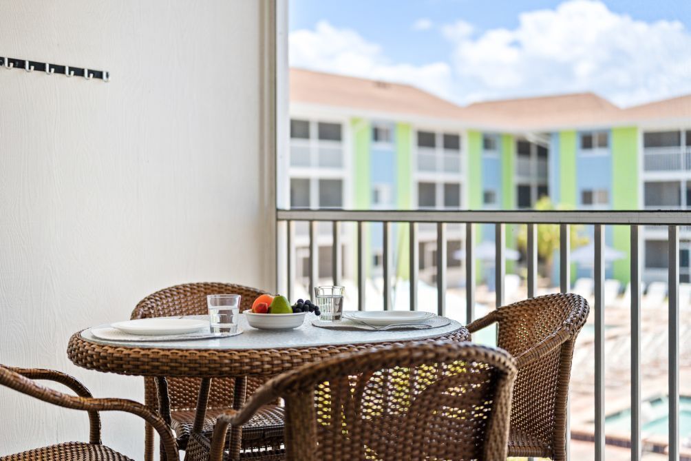 Een balkon met rieten stoelen, een ronde tafel en uitzicht op een gebouw in groen en wit.