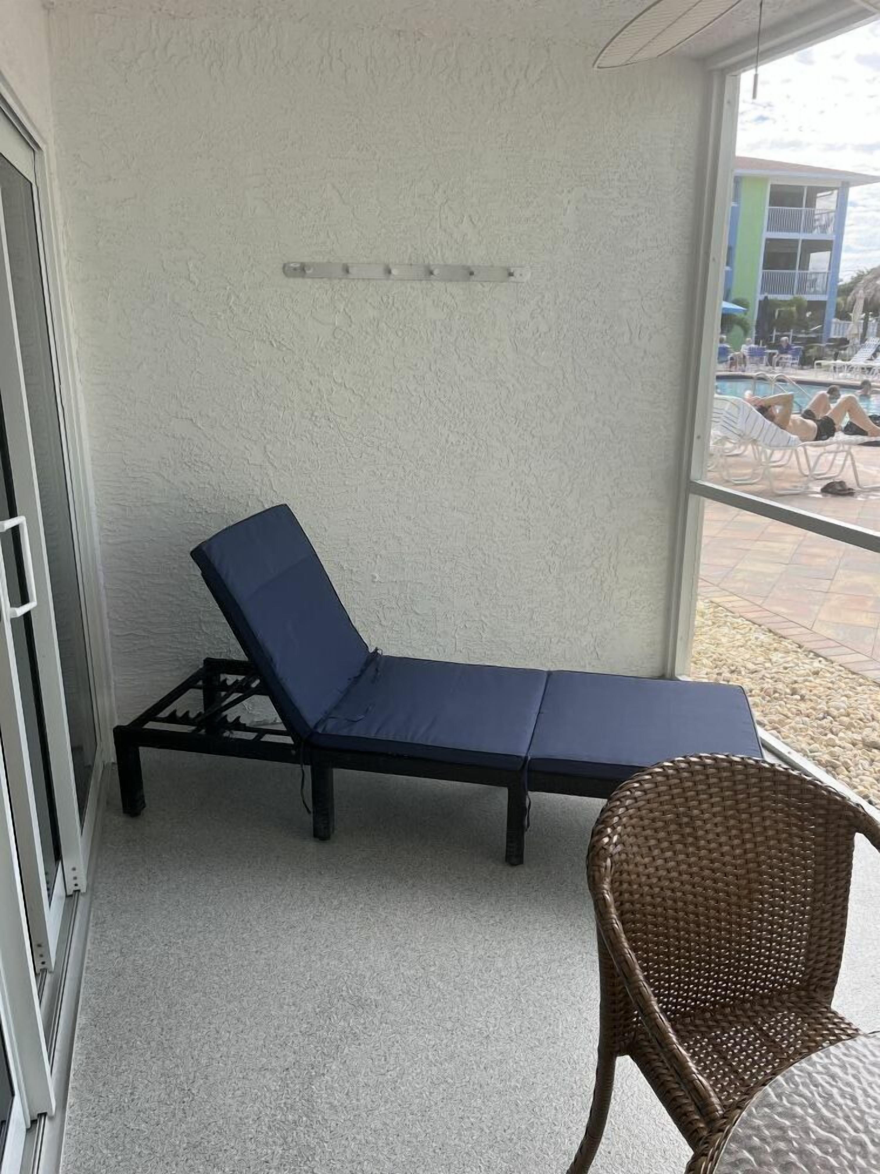 A sun lounger with a blue cushion and a wicker chair in a room with a textured white wall and a window overlooking a pool area.