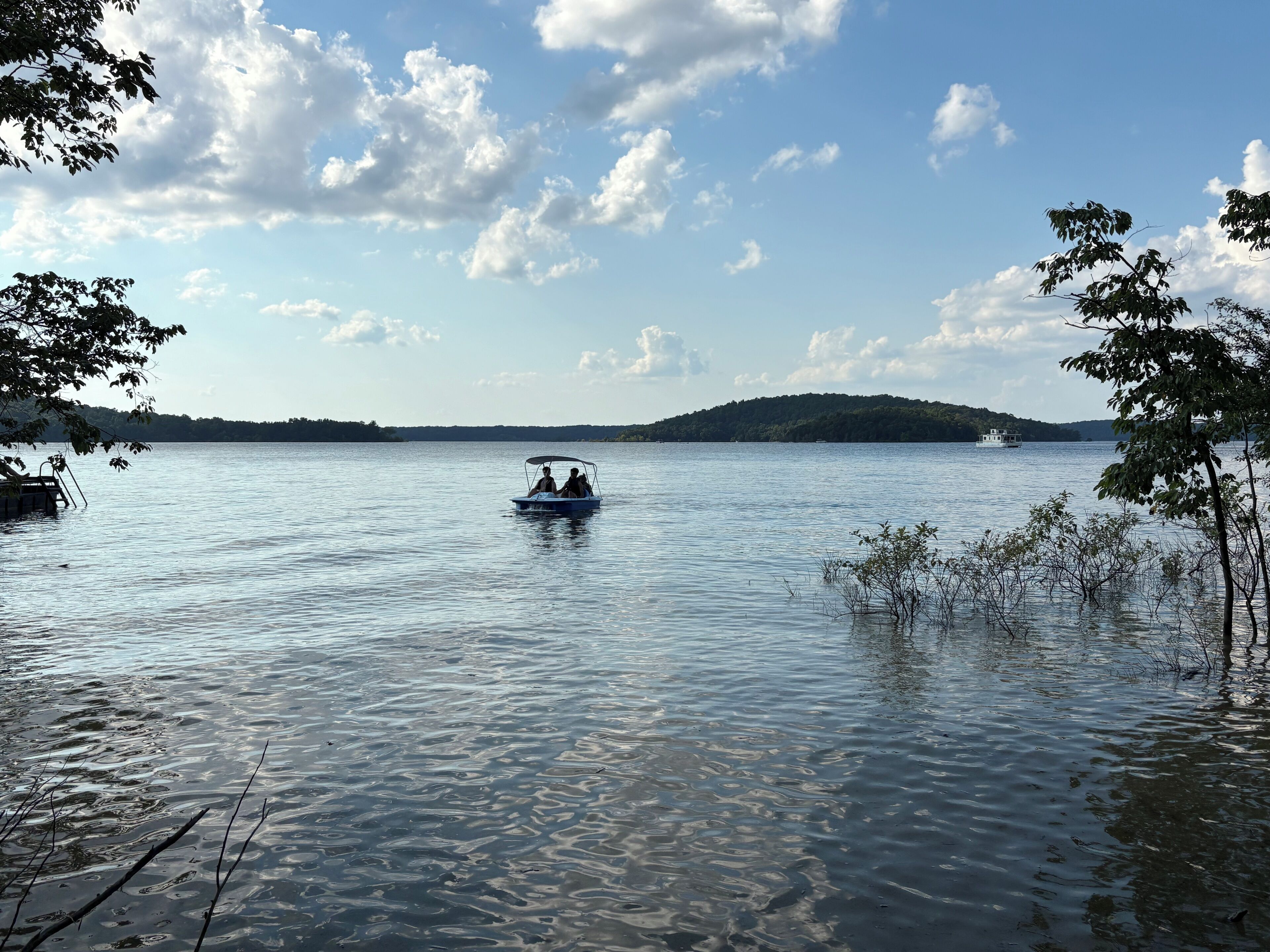 A calm lake with a boat in the center, surrounded by trees and a distant hill.