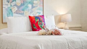 Un lit avec une literie blanche, un coussin coloré et un plateau avec des verres et des fleurs.