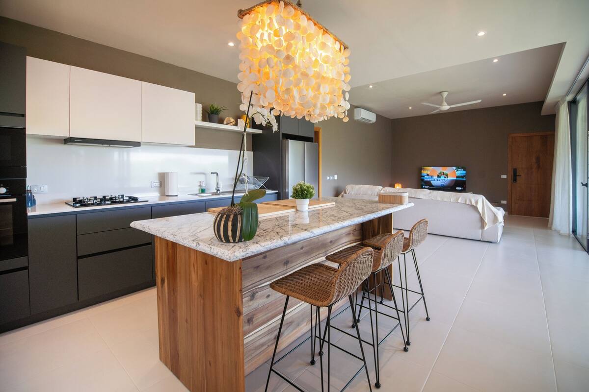 Una cocina moderna con una isla central, taburetes de bar, un televisor y un ventilador de techo.