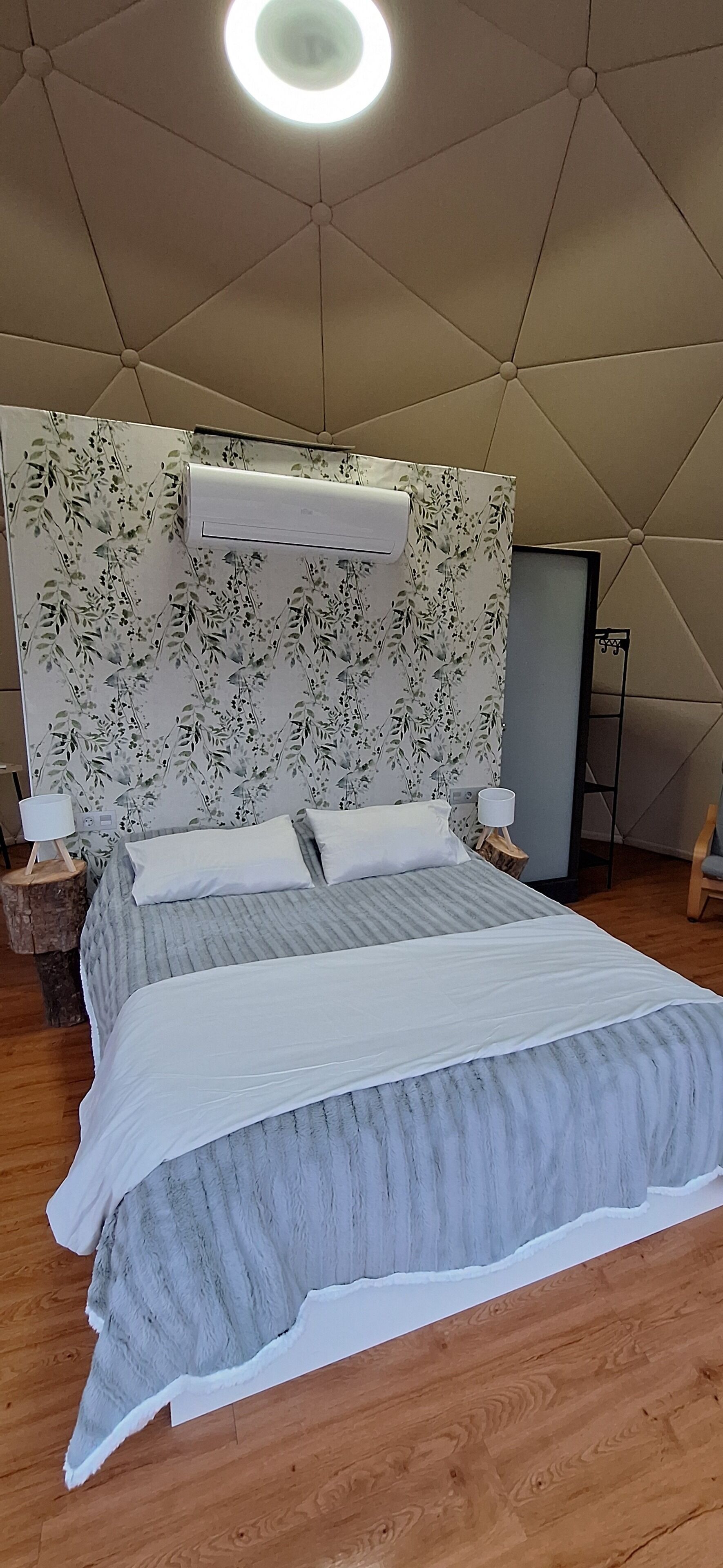 A bedroom with a large bed, a floral-patterned headboard, a ceiling light, and a nightstand with a lamp.