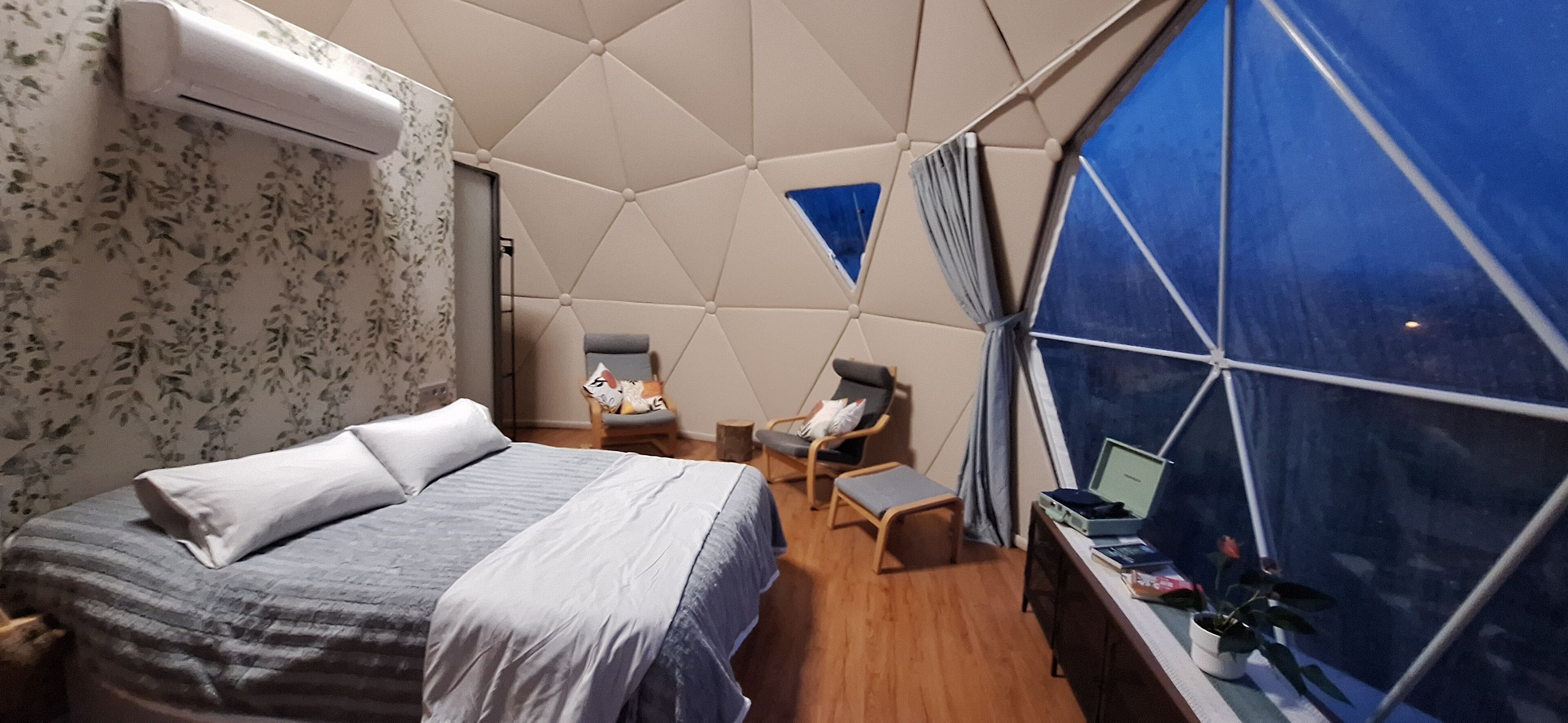 A modern room with a geometric ceiling, a bed with a grey and white duvet, a small desk with a laptop, and a window with a view.