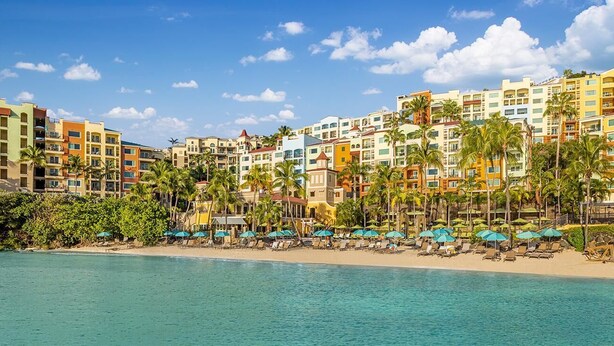 Un hotel frente a la playa con varios edificios de colores, palmeras y una playa de arena con sombrillas.