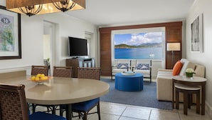 Una sala moderna con una mesa de comedor redonda, un reposapiés azul y un sofá con un cojín naranja. Un televisor está montado en la pared, y un ventanal ofrece vistas al mar.