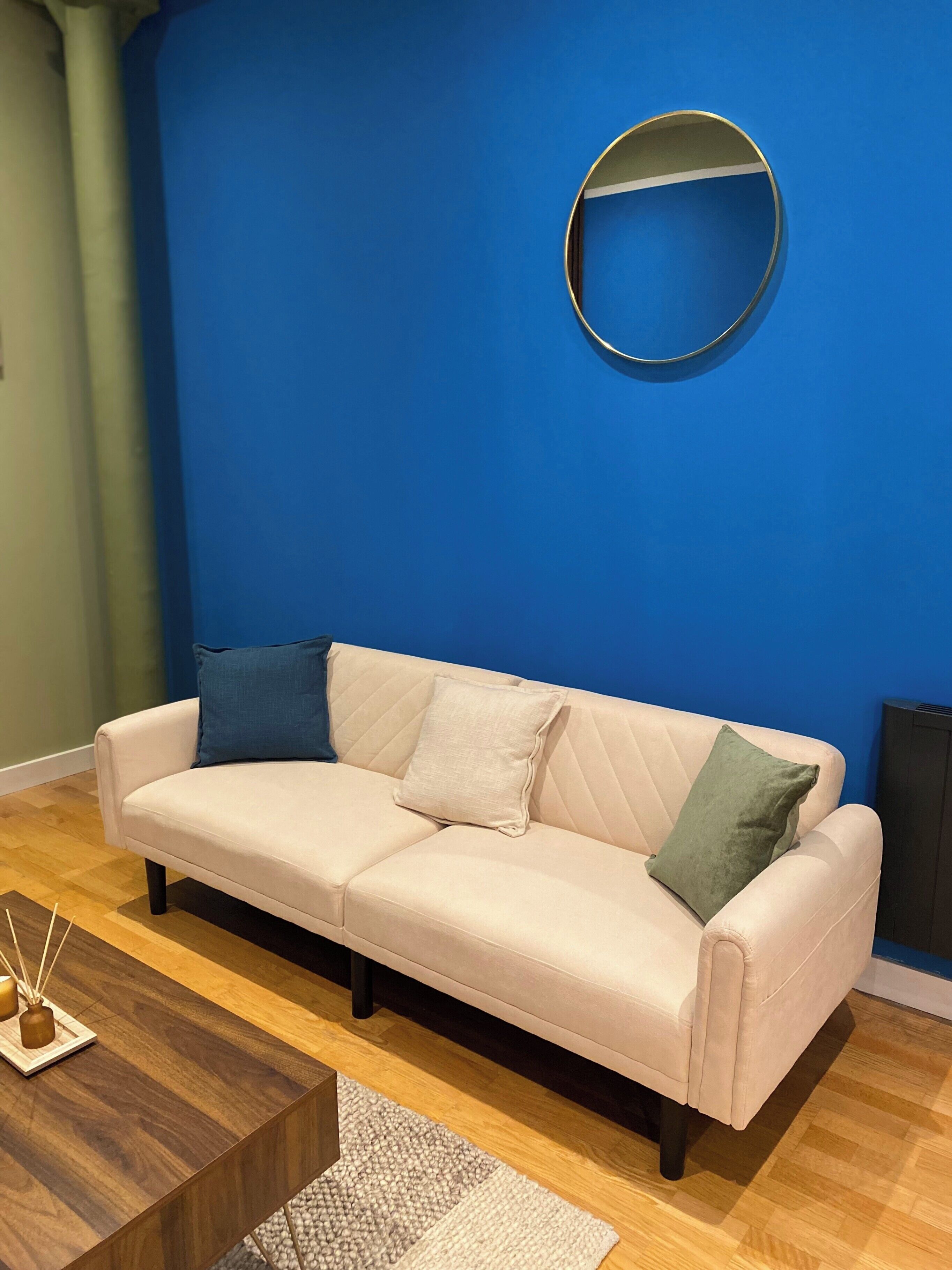 Un sofá beige con dos almohadas, un espejo redondo y una mesa de centro de madera en una habitación con pared azul y suelo de madera.