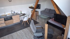 A modern kitchen with white cabinets, a dining area with a glass table and white chairs, a gray sofa, and a TV mounted on a wooden beam. - Appartement mit Dachterrasse, Monkebude (Monkebude)