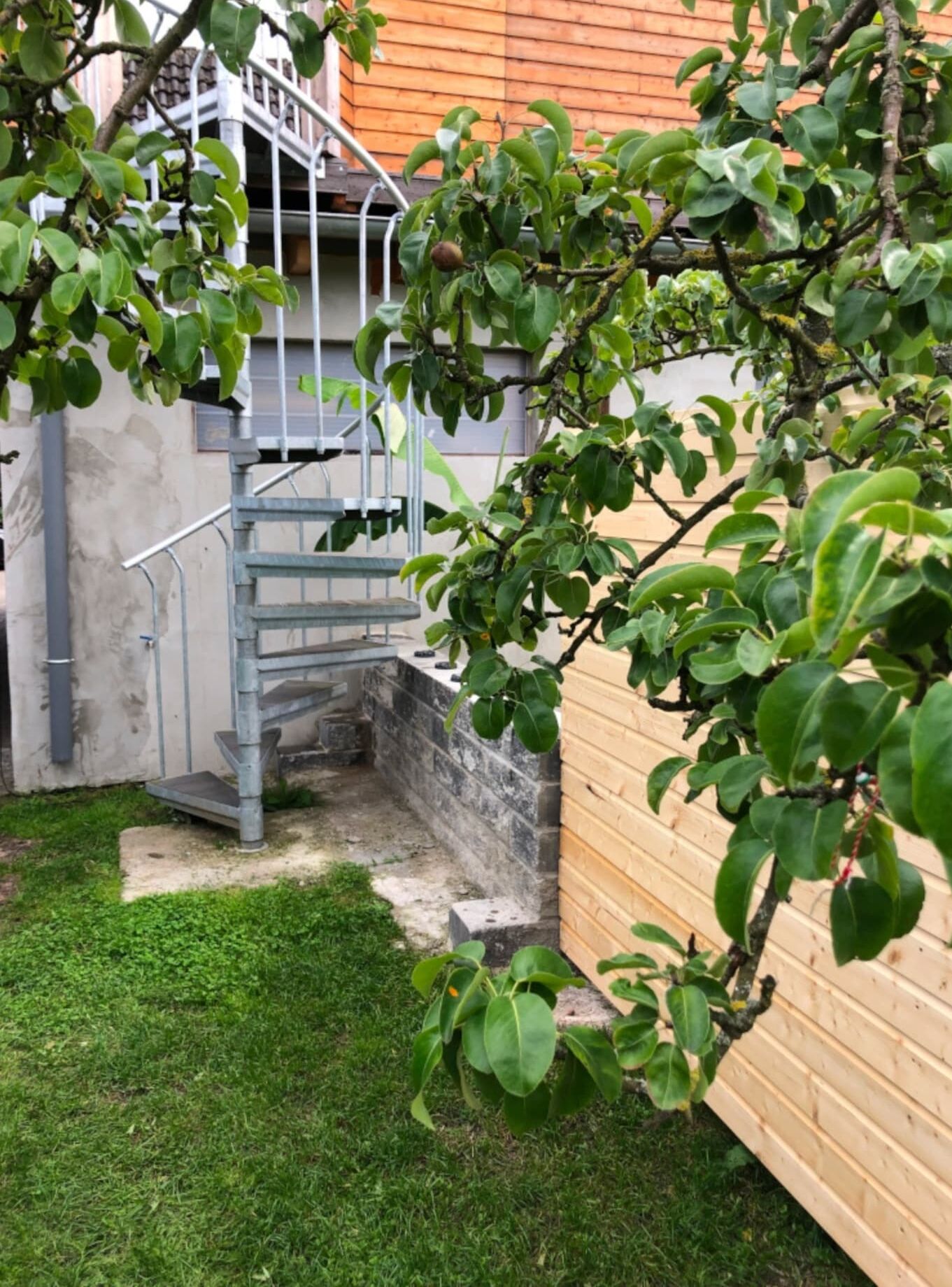 A small outdoor area with a metal staircase leading to a stone platform, surrounded by a wooden fence and a tree.