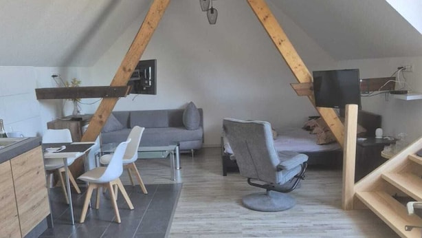 A modern loft-style room with a sloped ceiling, a dining area with a wooden table and chairs, a gray sofa, and a television. - Appartement mit Dachterrasse, Monkebude (Monkebude)