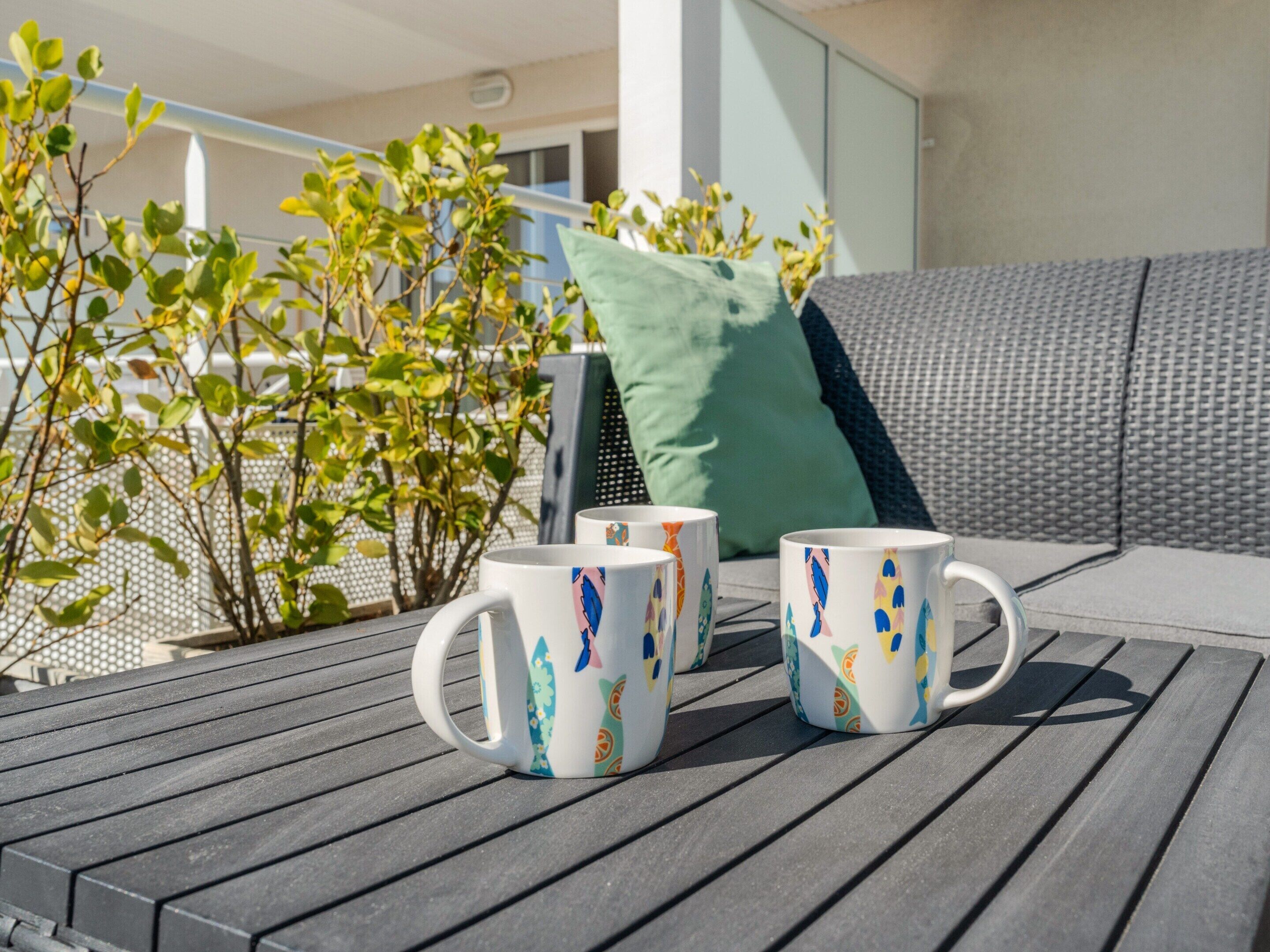 Una terraza de madera con tres tazas blancas decoradas con motivos coloridos, un cojín verde y un sofá gris.
