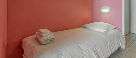 Un lit bien fait, avec du linge de lit blanc et une serviette pliée posée sur le côté, une lampe fixée au mur et un mur peint en rose.