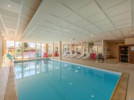 An indoor swimming pool with clear glass walls, multiple lounge chairs, and a tiled ceiling.