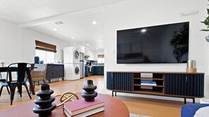 Un salon moderne avec un grand téléviseur à écran plat, une table basse ronde agrémentée de livres et d’objets décoratifs, un canapé et une cuisine en arrière-plan.