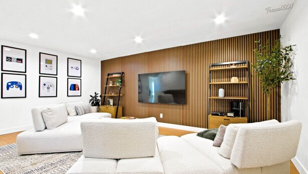 Un salon moderne avec un canapé d’angle, un téléviseur à écran plat, un lambris en bois et des manettes de jeux vidéo encadrées accrochées au mur.