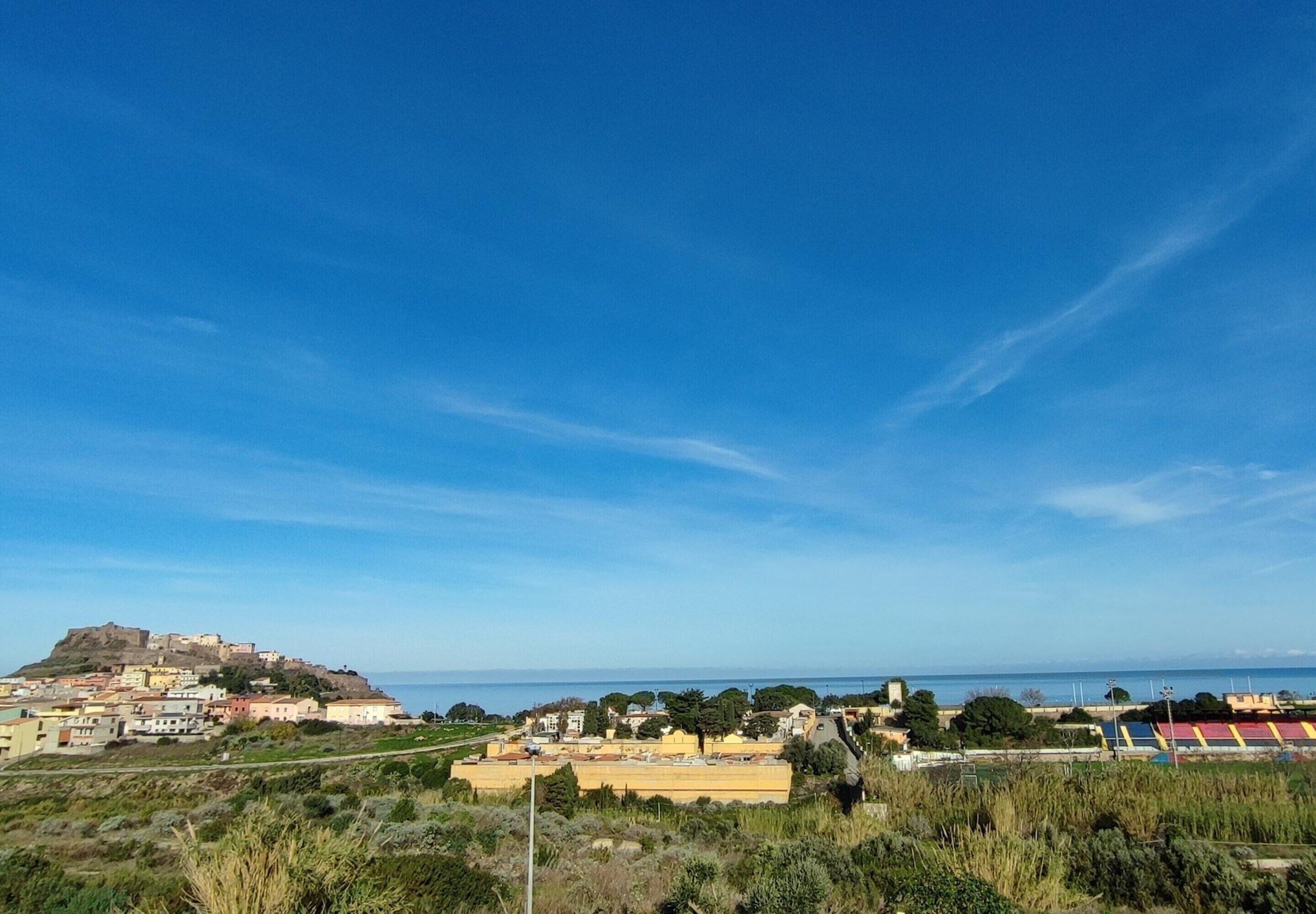 A coastal town with a prominent hilltop fortress, a mix of residential and commercial buildings, and a clear blue sky.