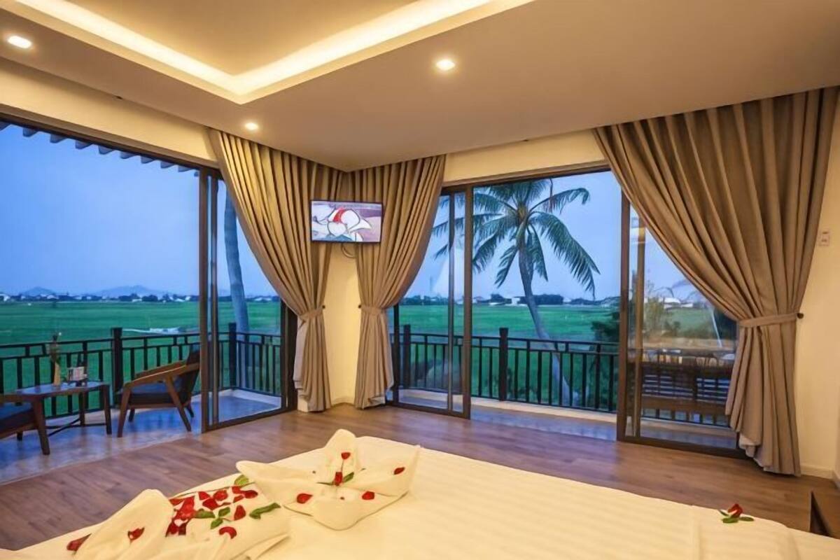 A hotel room with a bed, a TV, a balcony with a view of the countryside, and a dining area.