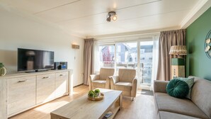 Un salon moderne avec un téléviseur à écran plat, une table basse en bois, des canapés beige et une grande fenêtre avec des rideaux.