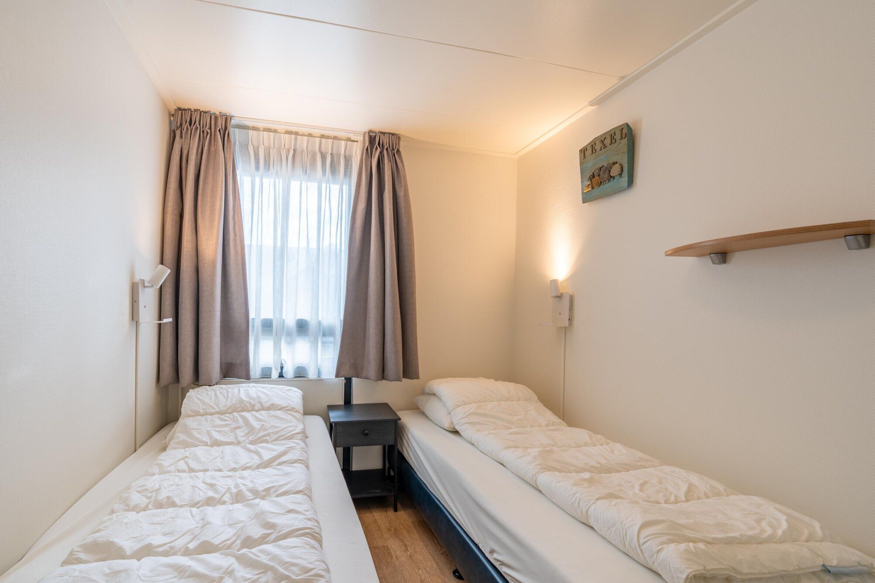 A small, clean room with two single beds, a small table, a nightstand, and a window with curtains.