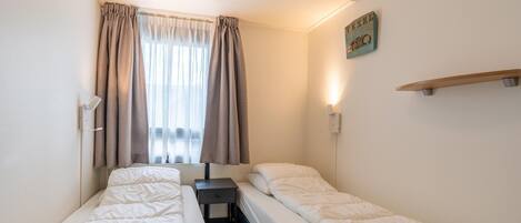 A small, clean room with two single beds, a small table, a nightstand, and a window with curtains.