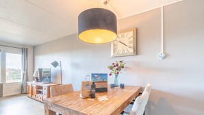 A dining area with a wooden table, chairs, a black lamp, a clock, and a vase with flowers. - Comfortable 4-person Apartment With Terrace and View (De Koog)