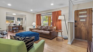Een moderne woonkamer met een bruine leren bank, een blauwe poef en een groene fauteuil. Er is een houten bureau met een laptop, een boekenkast en een lamp. De kamer heeft houten vloeren, een groot raam met oranje gordijnen en een houten deur met een plaquette.