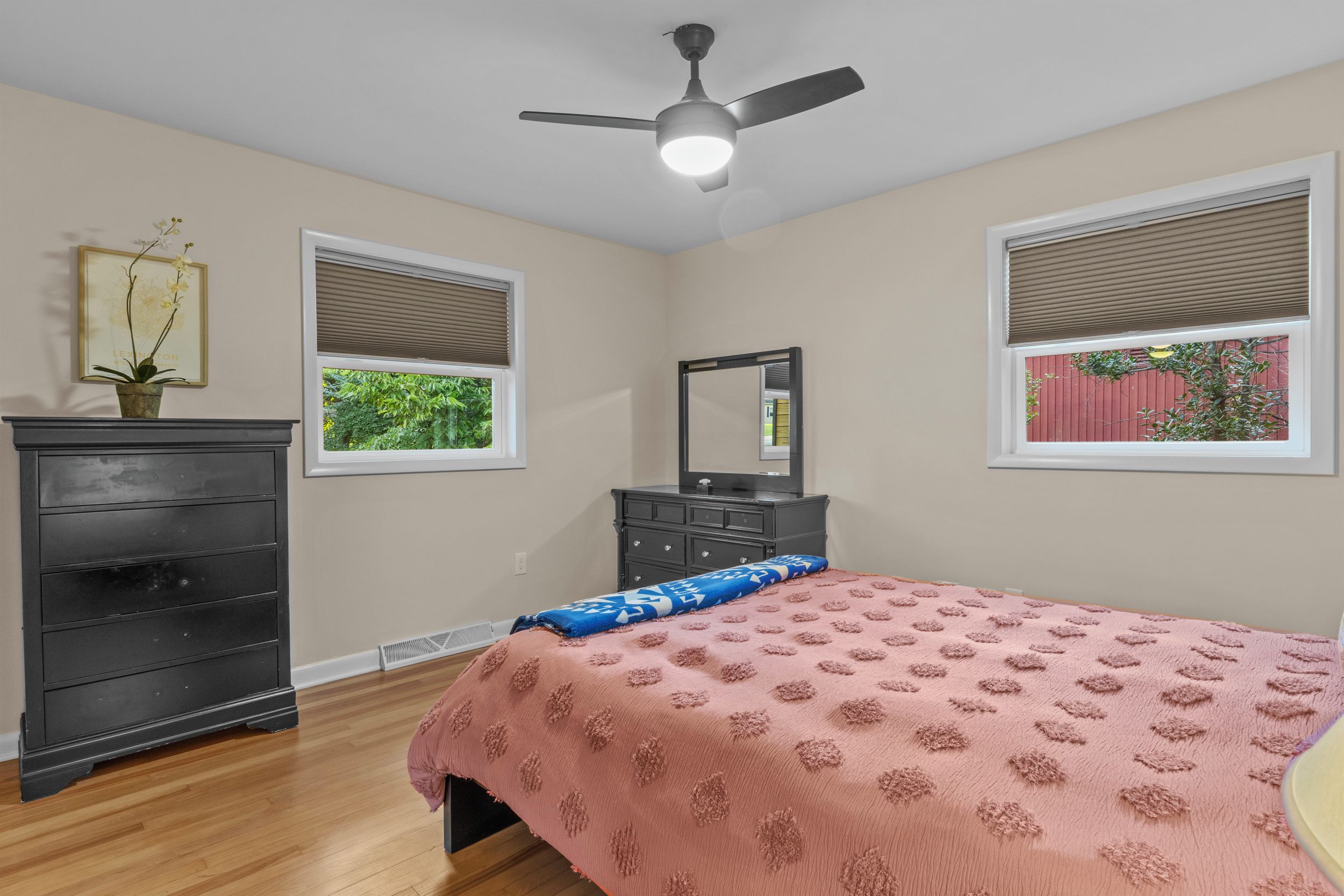 Une chambre à coucher comprenant un lit, une commode, un miroir, un ventilateur de plafond et des fenêtres.