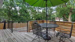 Een houten terras met een ronde tafel en stoelen, een groene parasol en een houten hek.