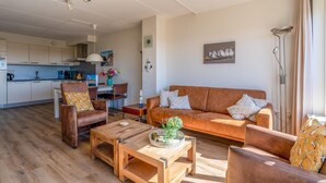 A modern living room with a sofa, armchairs, and a coffee table. There is a kitchen in the background with cabinets and a dining area. - 4-person Apartment With Island-sea View (De Koog)