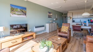 A living room with a TV, wooden furniture, a dining area with a table and chairs, and a kitchen in the background. - 4-person Apartment With Island-sea View (De Koog)