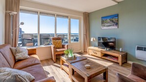 A spacious living room with a large window, a sofa, a chair, a coffee table, a TV, and a painting on the wall. - 4-person Apartment With Island-sea View (De Koog)