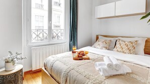 A neatly arranged hotel room with a large bed, a small side table with a plant, a window with a view of buildings, and a tray with croissants and juice. - Cozy 1BR in Montorgueil (Paris)