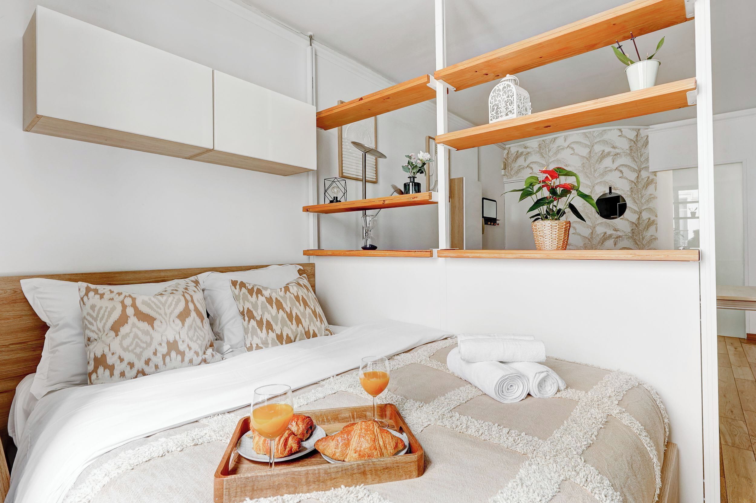 Una cama bien hecha con una bandeja de croissants y jugo, una mesita de noche con una lámpara, y una estantería con objetos decorativos.
