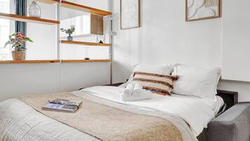 A neatly arranged bedroom with a bed, a bedside table, a lamp, a window with curtains, and a wall-mounted shelf with decorative items.