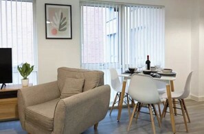 Ein modernes Wohnzimmer mit einem beigefarbenen Sofa, einem Esstisch mit weißen Stühlen und einer Zimmerpflanze.