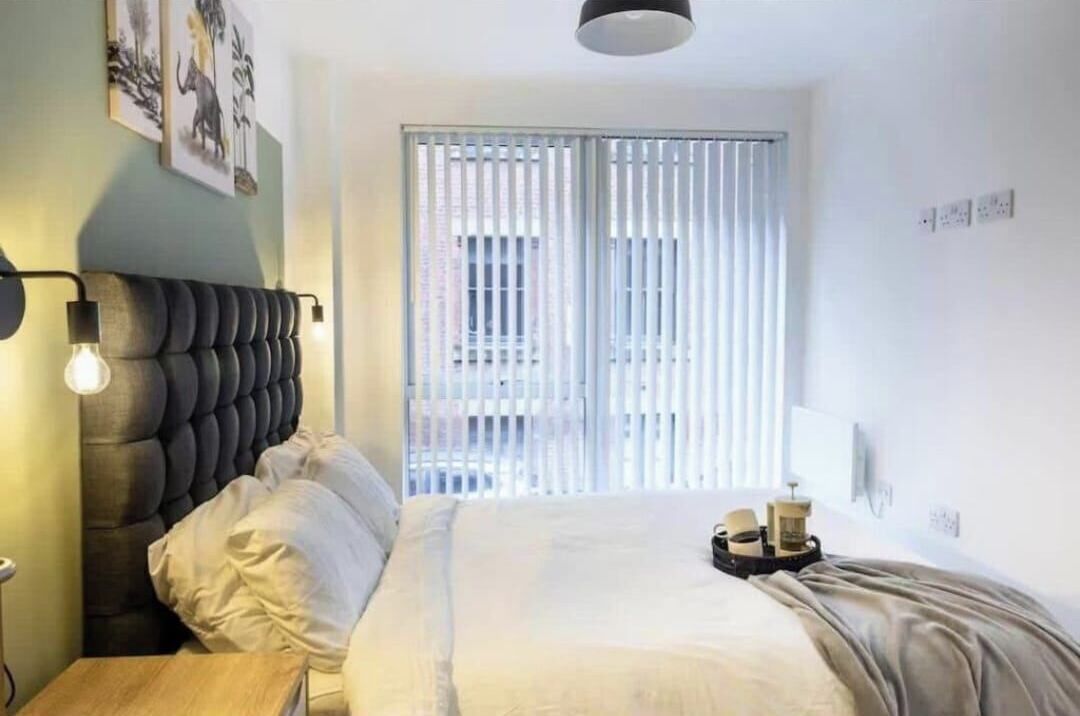 A bedroom with a tufted headboard, a bed with white linens, a bedside table with a lamp, and a window with blinds.
