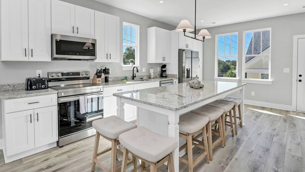 Cozinha moderna com armários brancos, eletrodomésticos de aço inoxidável, uma ilha grande com banquetas e janelas amplas que proporcionam luz natural.