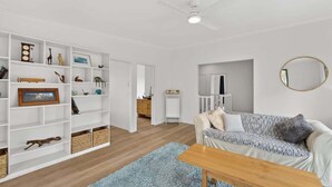 A living room with a white sofa, a wooden coffee table, and a bookshelf with various items. - Kareelah | Ocean Views & Walk to Beach (Guerilla Bay)