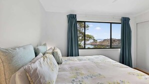 A bedroom with a large bed, a window with curtains, and a view of the outdoors. - Kareelah | Ocean Views & Walk to Beach (Guerilla Bay)